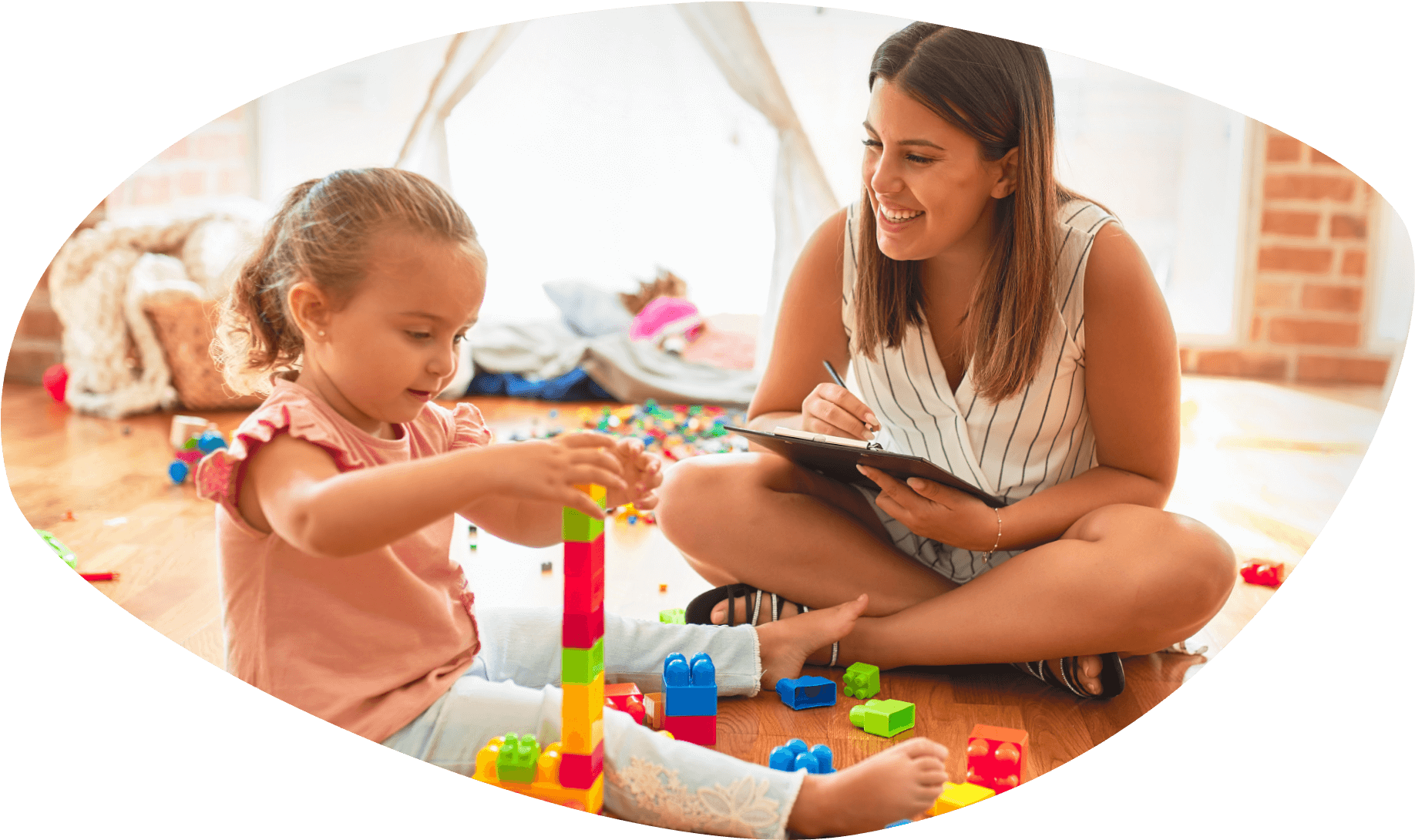 Girl sitting on floor is building a block tower while young woman sitting on floor takes notes