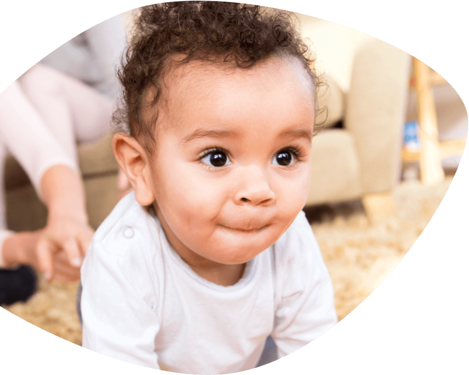 A small child is pulling herself up and looking ahead intently while a protective adult sits nearby